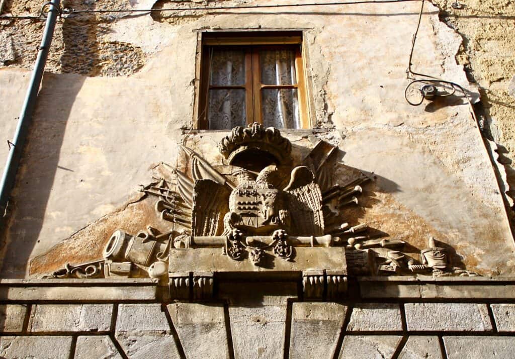 Detailed stone sculpture on a weathered building wall with a window above, showcasing Abruzzo's historic charm.