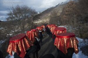 People carry decorated tables on their heads during a cultural festival in Abruzzo, against snowy mountains and leafless trees.